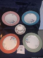 Four Aynsley dessert plates with different pastel colored rims (pink, blue, orange, green) and single tea cup with matching saucer placed on black cloth background