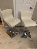 Two white swivel bar stools with chrome square bases against a white door and beige walls.