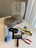 Photo showing entire dog care lot including the grey automatic water dispenser with transparent bone-patterned container, electric training collar with strap, nail clippers, grooming scissors, slicker brush, and portable water bottle dispenser arranged on a white table.