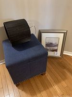 Blue fabric ottoman with brown fabric lampshade resting on top, framed polar bear print leaning against wall on floor.