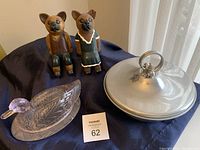 Photo showing two wooden cat figurines, a glass duck-shaped butter dish, and a silver plated casserole dish with lid on a black cloth