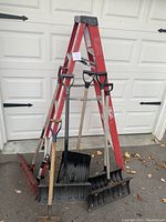 Red metal A-frame ladder and five shovels arranged in front of garage door on paved driveway.