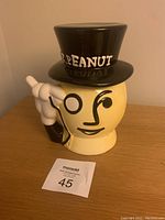 Front view of Mr. Peanut ceramic jar showing face with monocle and pointing hand, black top hat lid with word 'PEANUT' in white.