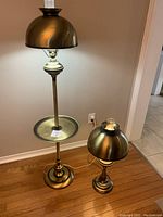 Both vintage brass hurricane lamps shown standing on hardwood floor, a tall lamp with tray and smaller lamp with dome shade.