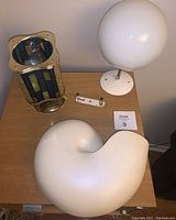Overview of three lighting pieces on wooden surface: stained glass brass lantern, white glass globe lamp, and white ceramic wall sconce in shell shape.
