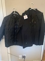 Two men's winter coats hung on hangers on a door, one quilted puffer and one softshell.