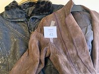 Close-up of both the brown leather jacket and the black ski suit hung next to each other, showing jacket collar with patterned lining and ski suit fabric details.