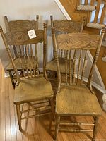 Four wooden dining chairs with carved cameo-style backs, one chair missing a spindle on backrest, sitting on hardwood floor near a staircase