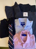 Photo of three pairs of youth boys dress pants in dark colors, two long sleeve shirts (blue and pink) folded, a black sweater vest and a striped tie.