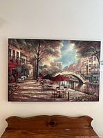 Framed wall painting showing sidewalk cafe tables, chairs, trees, canal and city buildings in warm tones.
