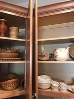 Cupboard shelves showing woven baskets, wooden bowl, small ceramic bowls and large floral ceramic teapot without lid.