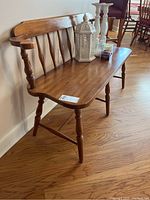 Wooden bench with spindle back and armrests, sitting on hardwood floor against white wall with some decor items on seat.