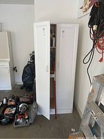 White tall cabinet with two doors, doors partially open revealing inside shelving. Cabinet located in garage with tools and ladder nearby.