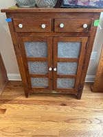 Full front view of wooden cabinet showing two drawers with white knobs and two lower doors with decorative punched tin panels.
