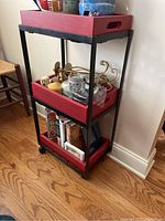 Side angle of the metal cart showing three red wooden box shelves containing miscellaneous items like kitchen utensils and books, on hardwood floor.
