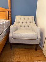 Front view of single button tufted upholstered armchair next to wooden bed frame on hardwood floor under blue and white wall.