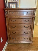 Front view of wooden chest of drawers showing five drawers with ornate metallic handles and carved embellishments on corners, highlighting wood grain and condition.