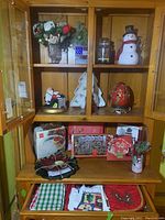 Wide view of shelf with various Christmas decorations including floral arrangements, snowman figure, platters, and linens