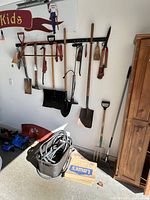 Wide shot showing various yard tools hanging on wall rack including shovels, pick axe, saw, crow bar, pruning shears, and a basket with garden hose coiled inside.