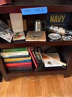 Shelf showing military memorabilia including pins with ribbons in a case, a pewter cup, metal plate, a ship model in a bottle, coins, and several books beneath.