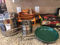Overview photo showing large stockpot, double boiler, frying pan, Romertopf German clay Dutch oven with box and cookbook, and Lodge red enamel Dutch oven in box.
