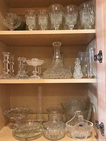 Six shelves of assorted clear glassware including crystal and pressed glass items such as candleholders, decanters, and wine glasses.