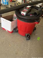 Craftsman red 12 gallon shop vacuum on wheels with coiled black hose and wand attachment, accessory items in white cardboard box nearby.