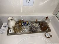 Mirrored vanity tray holding several decorative and functional items including white ceramic Lennox vase, lead crystal ring holder, glass dome, metal basket, and pocket watch on a bathroom countertop.