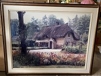 Framed photo print of a thatched cottage surrounded by flowers and greenery in a classic wood frame with cream matting.