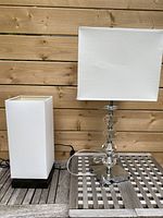Photo showing both lamps side by side on a wooden surface against a wooden background. The smaller lamp has a white shade and a wooden base; the taller lamp has a chrome and clear geometric base and a larger white shade.