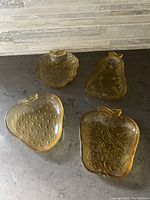 Four amber-colored glass appetizer plates shaped as an apple, pear, strawberry, and grapes, displayed on dark countertop.