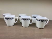 Front and side view showing all six white milk glass mugs with blue floral pattern near the rim and sturdy handles.