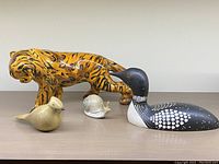 Four animal figurines shown together on a table: large orange ceramic tiger-striped cat, black and white carved Loon bird, small beige Cedar Waxwing bird, and small ceramic snail figurine.