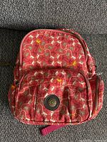 Top down view showing red backpack with colorful monkey print and front circular logo patch on zip pocket.