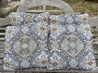 Four matching decorative throw cushions arranged on a curved wooden bench, showing the front patterned side with varied blues, beige, and browns in a symmetrical pattern.