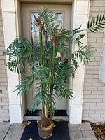 Full front view of the faux bamboo tree showing height, spread, multiple green stalks with brown fibrous tips, and green palm-like leaves. Standing on a doorstep.