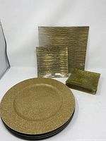 Photo showing 4 small square appetizer plates stacked, 2 large square serving plates behind, and 4 round chargers stacked in front, all gold-toned with various textures.