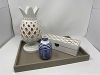 Photo showing all items: white ceramic pineapple candleholder, blue and white vase, white carved wood box with gold pattern, and taupe serving tray