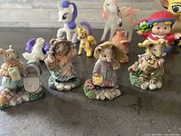 Photo showing four animal figurines dressed in clothes holding gardening and flower-themed accessories sitting on bases, with three small My Little Pony toys in background.