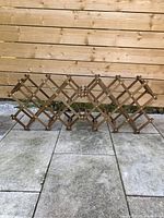 Two teak accordion wine racks set spread open on stone floor against wooden fence background.