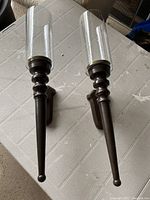Photo of two large matching metal candle sconces with attached clear glass cylindrical shades, showing the front view with details of metalwork and glass condition.