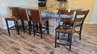 Five counter height bar stools in two sets next to a kitchen counter with stone facade, variegated granite countertop, and wooden floor.