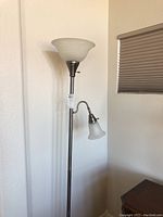 Full view of the metal floor lamp with two lamps and frosted glass shades, placed near a wall corner and window.