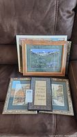 Six framed prints and photos stacked showing a variety of images and frames including mountain landscape, street scene, and decorative text art