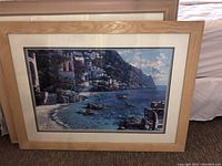 Three stacked framed prints showing coastal mountain village seascape with houses, water, and sky, natural wood frames, matting visible.