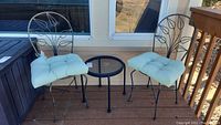 Two black metal outdoor chairs with floral/leaf design backs, beige cushions, and small black metal mesh side table set on outdoor deck.