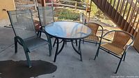 Full view of the patio furniture set outdoors on concrete patio including glass top round table and two sets of chairs.