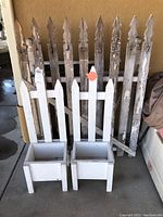 Photo showing two white wooden picket fence planter boxes with peeling white paint and three sections of old white picket fencing with heavy peeling and weathering.