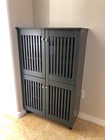 Front view of entire gray storage cabinet with four slatted doors and silver knobs, positioned in corner against beige wall and tan tiled floor.