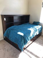 Twin bed shown fully assembled with bedding and pillows, featuring storage drawers under the bed and a bookcase-style headboard with shelves.